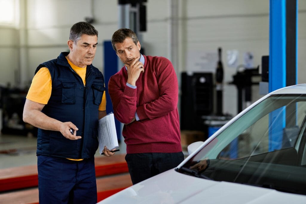 Two men standing next to a white car in an auto body shop garage.